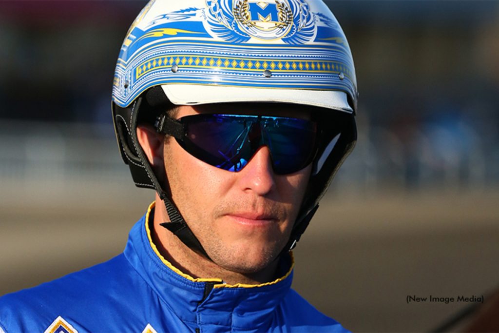 Close-up of a harness racing driver James MacDonald wearing a decorated helmet, sunglasses, and blue racing silks